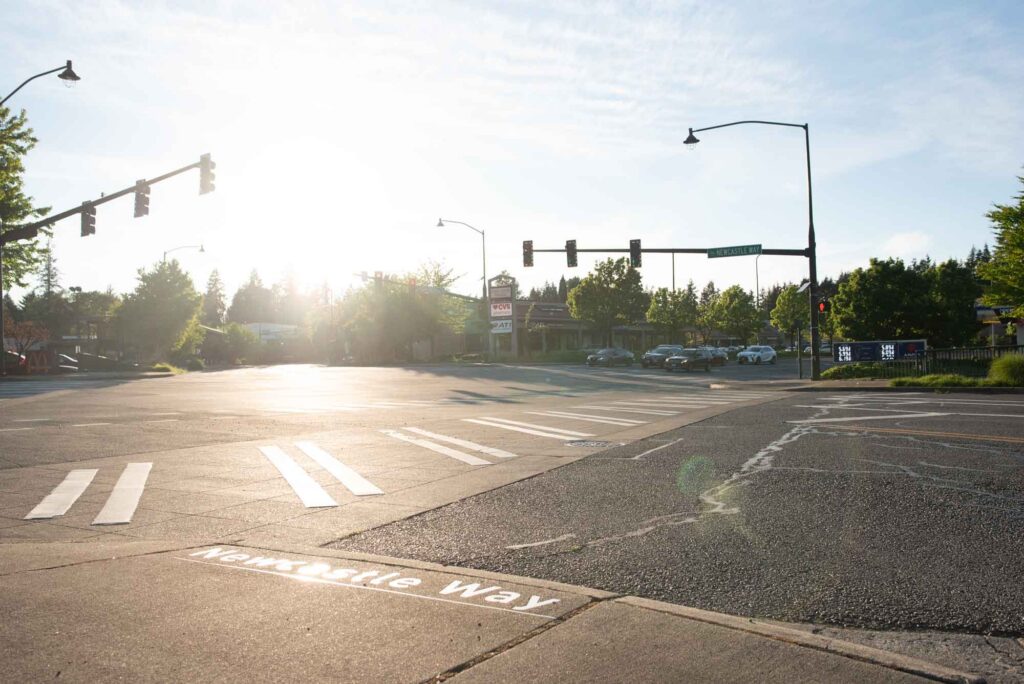 The intersection of Newcastle Way and Coal Creek Parkway in Newcastle Washington. Newcastle Way is visible on the sidewalk. Cars are stopped at a stoplight and the crosswalk is visible.