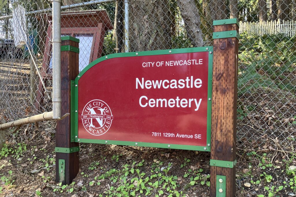 A red and green entrance sign for Newcastle Cemetery stands outdoors between two dark wooden posts. The sign reads “City of Newcastle – Newcastle Cemetery” and includes the City of Newcastle seal and the address “7811 129th Avenue SE.” Behind the sign is a chain-link fence with large trees and forested vegetation on a sloped hillside. Sunlight illuminates the sign and surrounding ground cover.